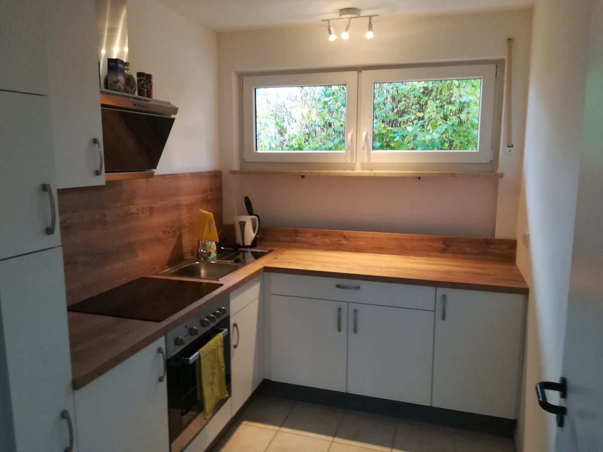 A modern kitchen features ample counter space with wooden surfaces and white cabinetry. A stovetop and oven are visible, alongside a kettle on the countertop. Natural light from two windows brightens the room, which includes a tiled floor and minimalistic accessories.