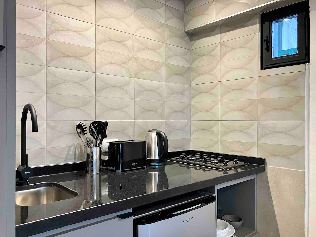 The kitchen area features sleek countertops and modern appliances, including a kettle and a coffee maker. Cooking utensils are neatly arranged in holders, while a small refrigerator sits beneath the counter. Natural light filters through a window, illuminating the textured tiles on the wall.