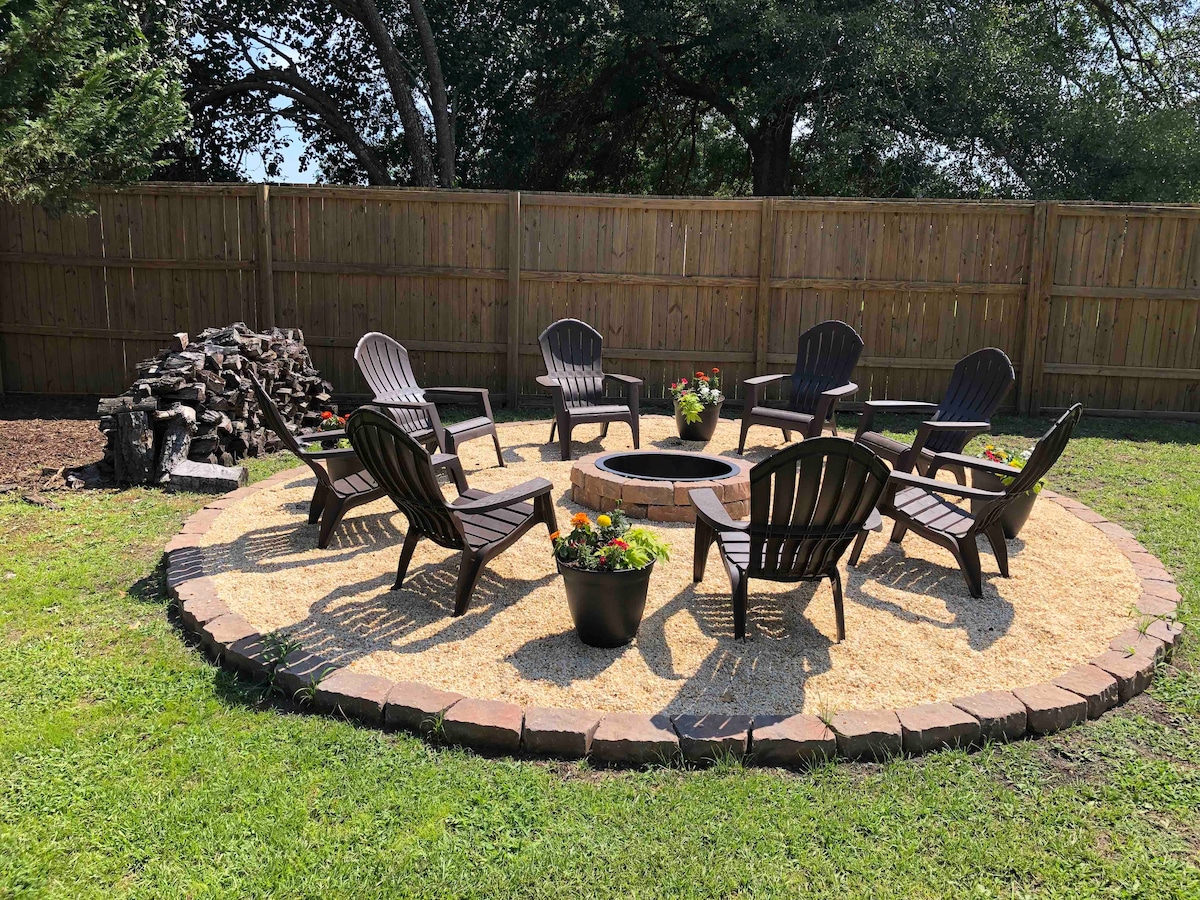 A firepit area is surrounded by several dark wooden chairs arranged in a circular pattern on a gravel surface. Flower pots add greenery, and firewood is stacked against a wooden privacy fence that encloses the space. The grass is well-maintained, creating a welcoming outdoor setting.
