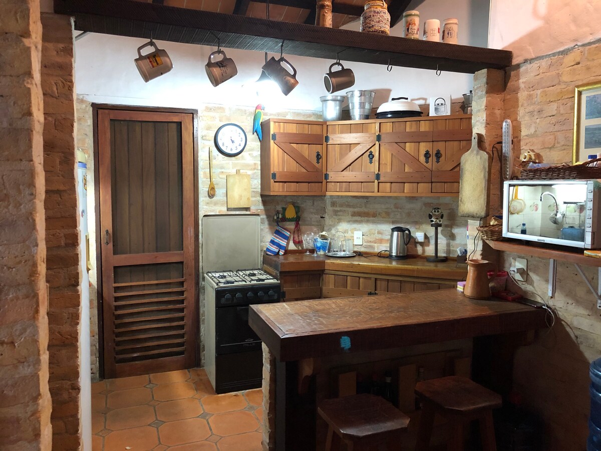 The kitchen area features rustic wooden cabinets with a variety of kitchenware displayed. A gas stove and a kettle are visible, along with a clock on the wall. A wooden bar table with stools offers space for casual dining, complemented by warm terracotta tiles.
