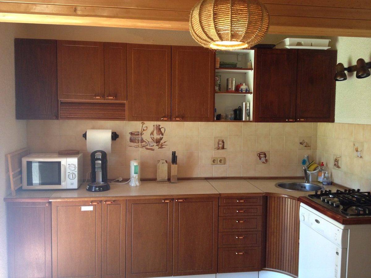 The kitchen features wooden cabinetry with ample storage, a gas stove, microwave, fridge, and dishwasher. A coffee maker is positioned on the countertop, complemented by beige tiled walls with subtle decorative touches. Natural light illuminates the space, enhancing its functional layout.