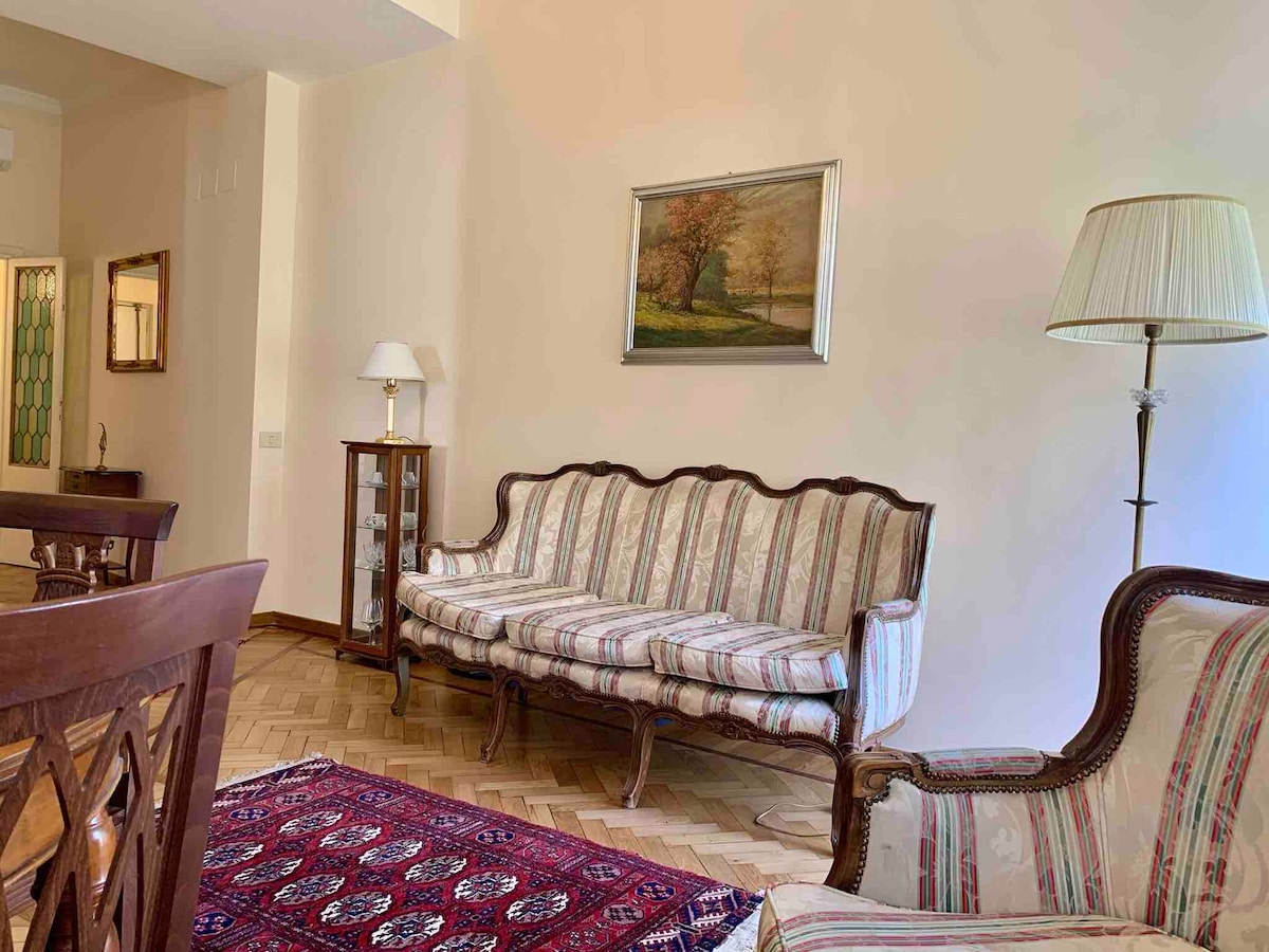 A seating area is presented with a vintage striped sofa and an accompanying armchair. A traditional Persian carpet covers the floor, and a framed landscape painting adorns the wall. A stylish floor lamp stands beside the sofa, enhancing the room's light and ambience.