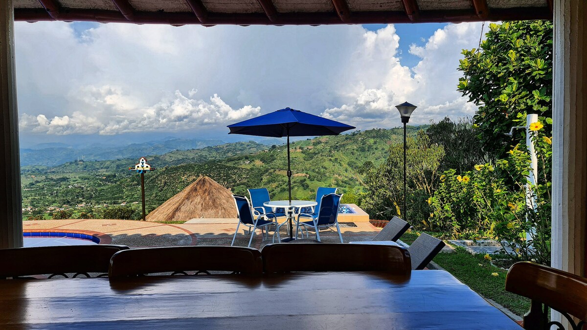 A scenic outdoor view is framed by a rustic wooden structure, showcasing a patio area with blue umbrellas and white lounge chairs. Lush green mountains and dramatic clouds create a picturesque backdrop, while vibrant flowers and a thatched-roof structure add to the tranquil setting.
