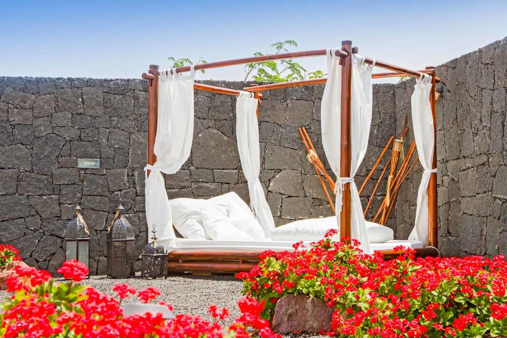 An outdoor lounging area is framed by a rustic stone wall, featuring a wooden frame draped with sheer fabric. Plush bedding invites relaxation, while vibrant red flowers surround the base, enhancing the tranquil setting.