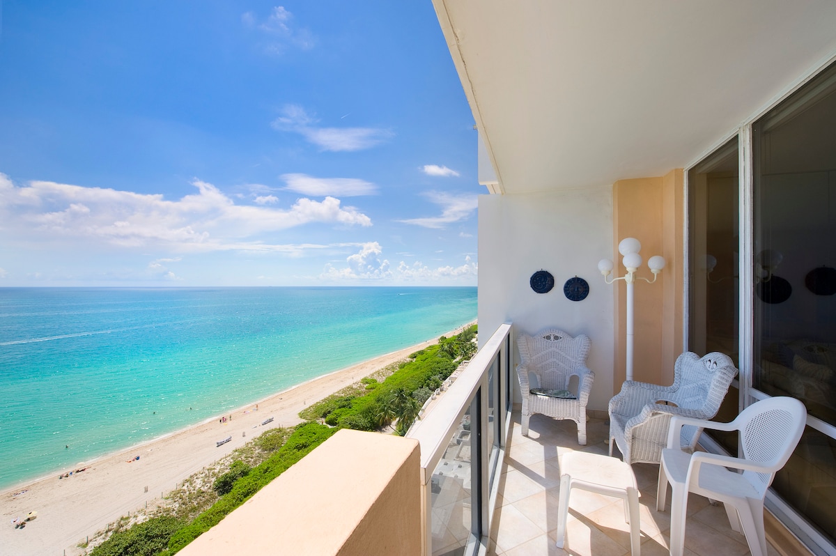 A balcony offers a panoramic view of the ocean and sandy beach, showcasing vibrant turquoise waters and a clear sky. Two white lounge chairs are placed on the balcony, along with a suitable table for enjoying the outdoors. The scene evokes a serene coastal atmosphere.