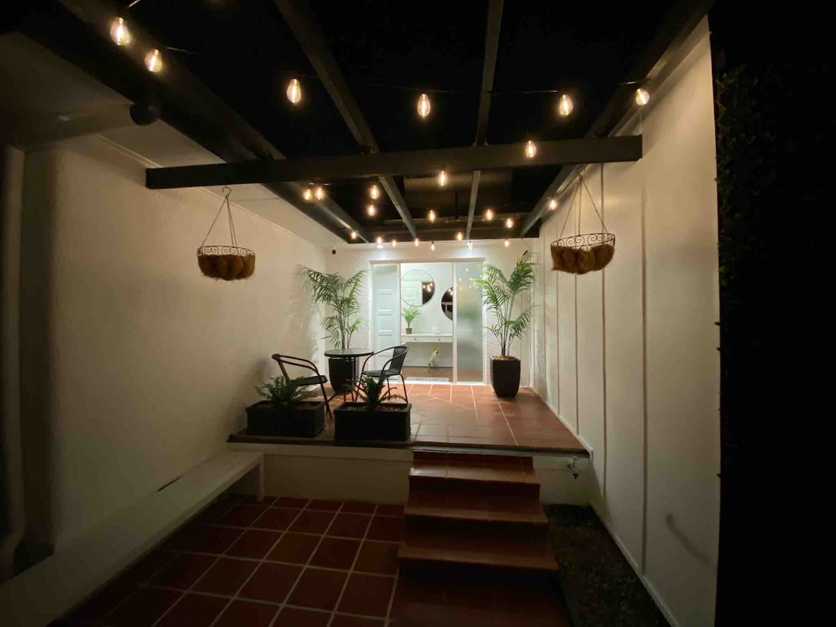 An outdoor area is presented with a tiled floor, featuring two chairs and hanging planters. Soft lighting from hanging bulbs creates a warm ambiance, while potted plants are placed around the space. The entrance is framed by white walls, leading to a visible interior.