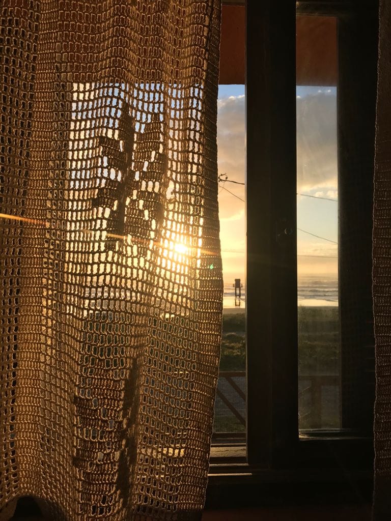 A glimpse through a lace curtain reveals a sunrise, casting warm light into the room. The sun's rays create a soft glow, highlighting the texture of the fabric and the window frame, while the ocean can be seen in the background.
