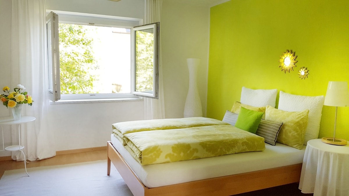A bright bedroom features a spacious bed with light green linens and decorative pillows against a vibrant green wall. A window opens outward, allowing natural light to fill the space, complemented by white curtains. A small table with flowers and a lamp adds a touch of warmth.