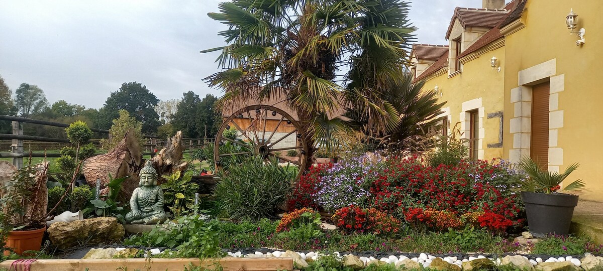 A vibrant garden is showcased, featuring a variety of plants including palm trees, colorful flowers, and sculptures. The well-maintained landscaping complements the warm yellow exterior of the nearby building, creating a serene outdoor space. A rustic wheel adds a charming touch to the setting.