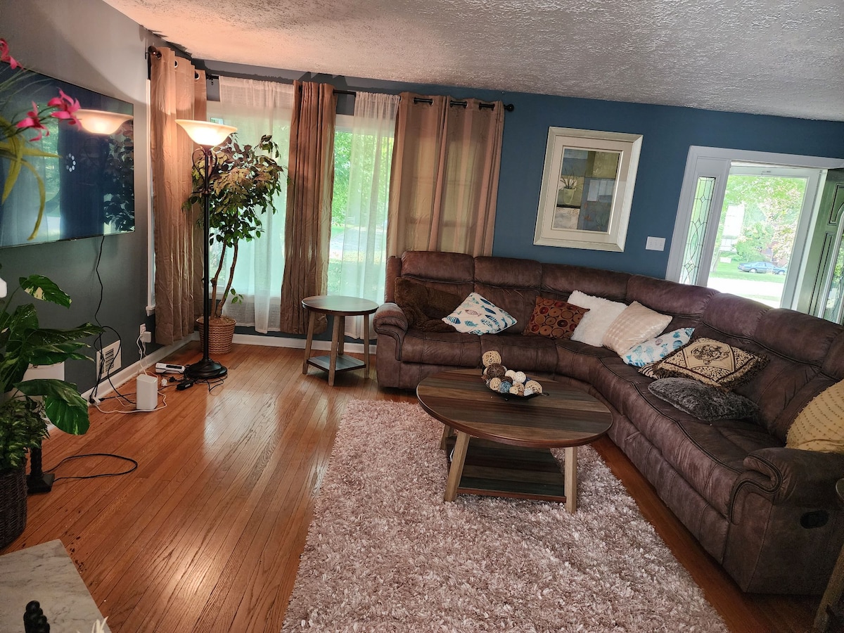 A spacious living area features a large sectional sofa adorned with decorative pillows. A round coffee table sits at the center, and a light rug adds softness to the hardwood floor. Natural light filters through sheer curtains, creating a bright ambiance.