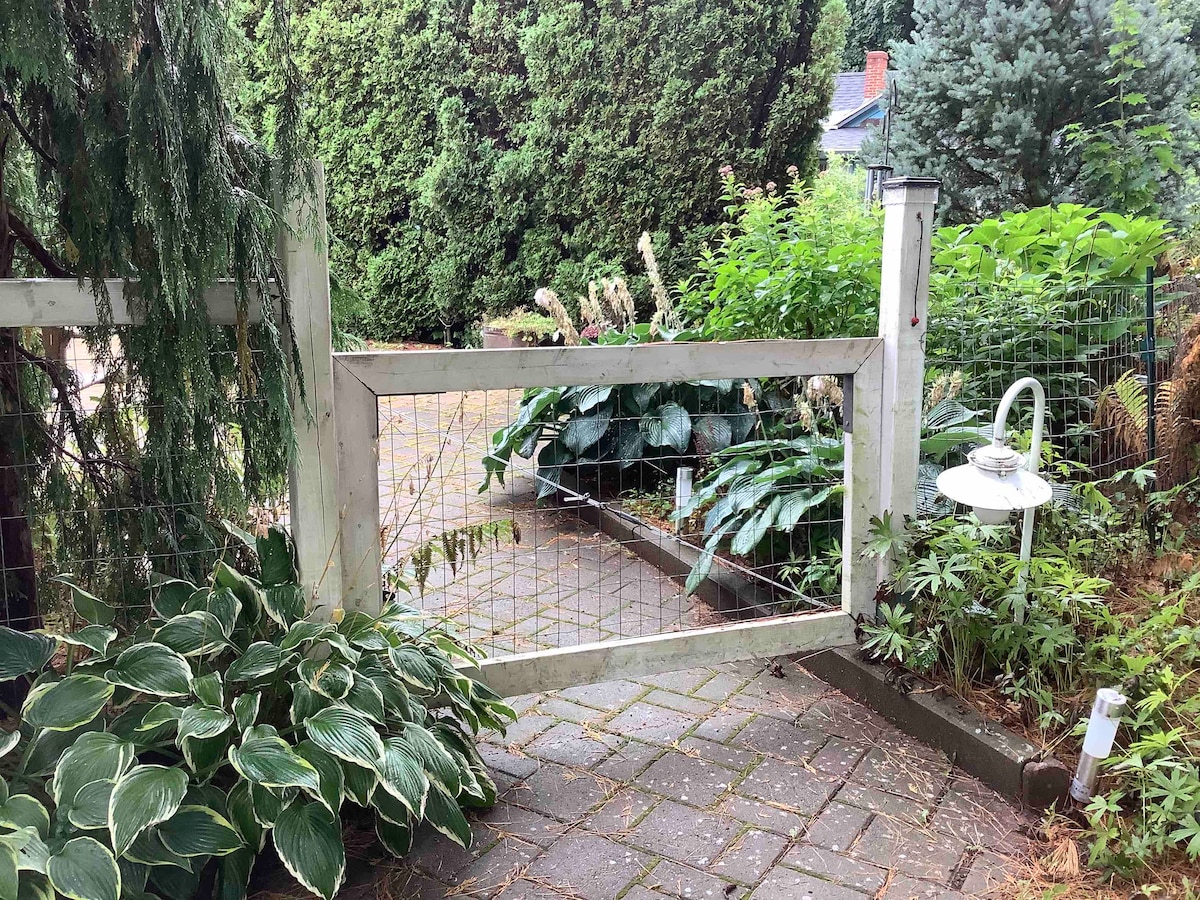 A garden entrance is framed by a simple white gate, set within a brick pathway. Lush greenery, including hostas and other plants, lines the walkway. A pathway light stands to the right, providing soft illumination amidst the vibrant foliage.