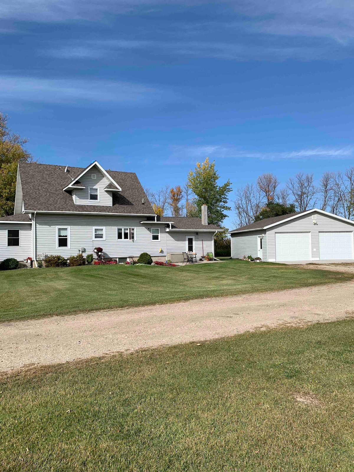 A charming two-story historic home is set amidst a lush green lawn. The house features a gabled roof and multiple windows, providing natural light. A spacious garage stands adjacent to the home, framed by a gravel driveway that leads to the entrance.