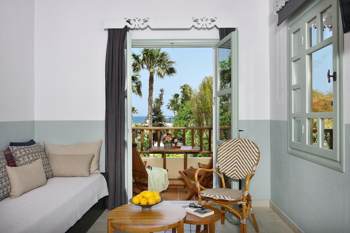 A cozy sitting area features a mix of comfortable seating, with a sofa complemented by decorative pillows. Wide doors open to a private balcony overlooking lush greenery and the sea, while natural light brightens the space through two framed windows.