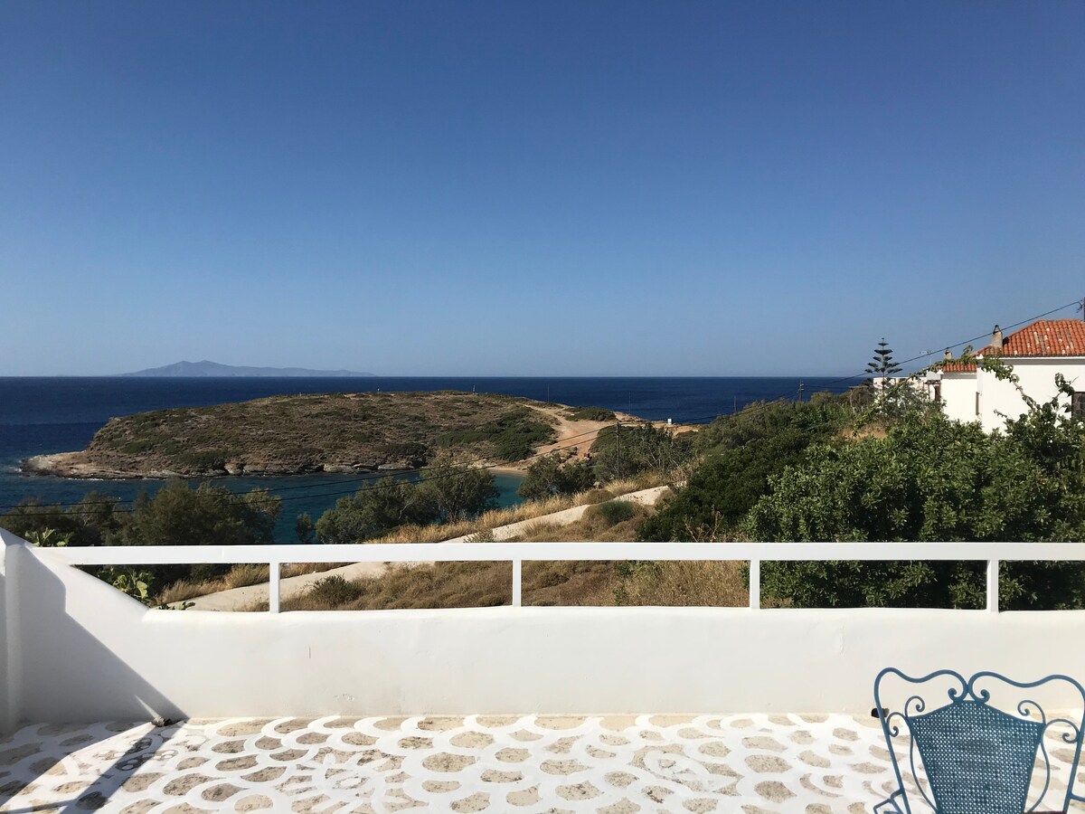 A spacious terrace offers a stunning view of the Aegean Sea and a nearby island. The surrounding landscape features dry, grassy terrain with scattered trees, while a clear blue sky enhances the serene atmosphere.