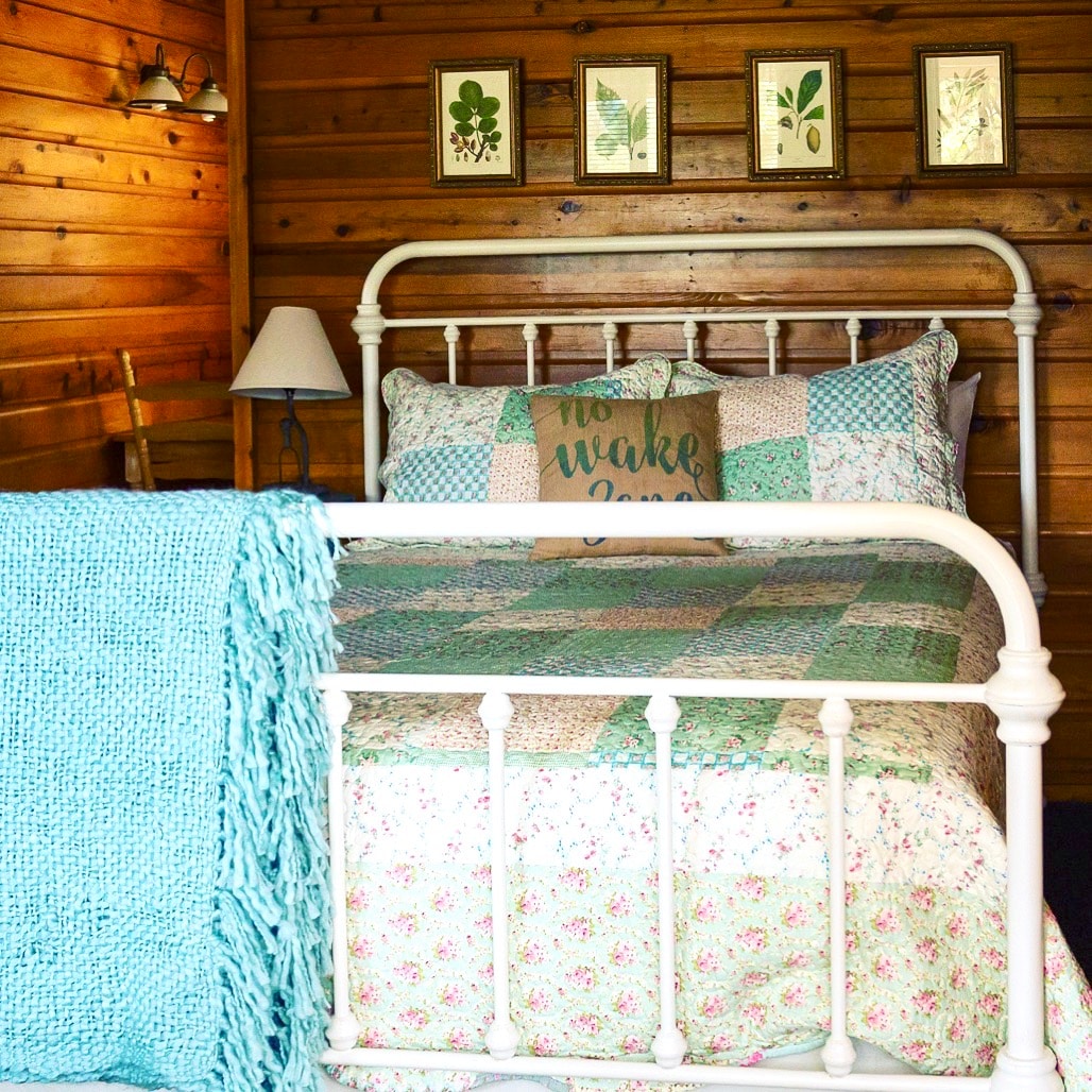 A cozy sleeping area features a white metal bed with a patchwork quilt in soft colors. Above the bed, framed botanical art adds a natural touch. A knitted throw in light turquoise drapes over the bed, and a bedside lamp provides soft lighting.