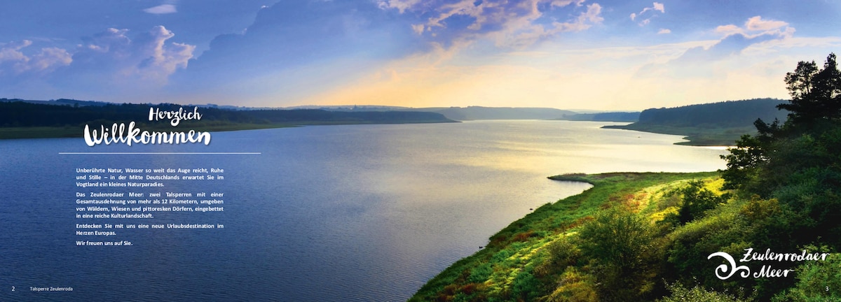 A serene view of a large lake is presented, framed by lush greenery along the shoreline. The sky reflects hues of blue and soft gold, hinting at sunrise or sunset. Gentle waves can be seen on the water's surface, enhancing the tranquil scene.