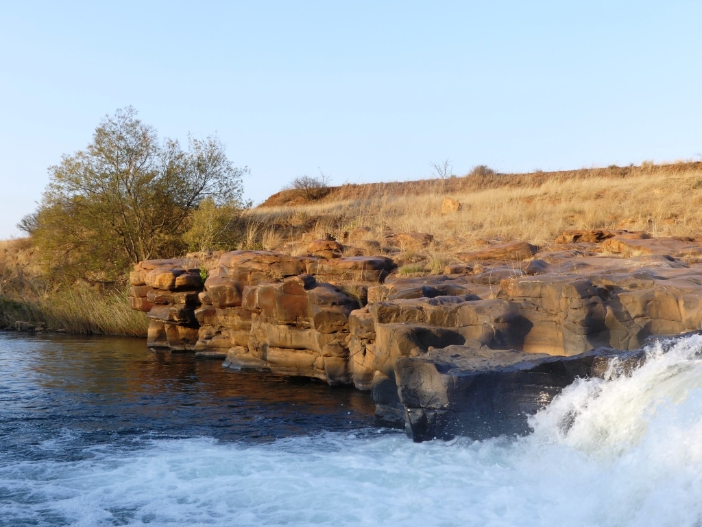 A rocky riverbank is presented, with textured, reddish-brown stones contrasting against the clear water of the river. A sparse tree grows near the edge, and golden grass is visible in the background along a gently sloping hill under a clear blue sky.