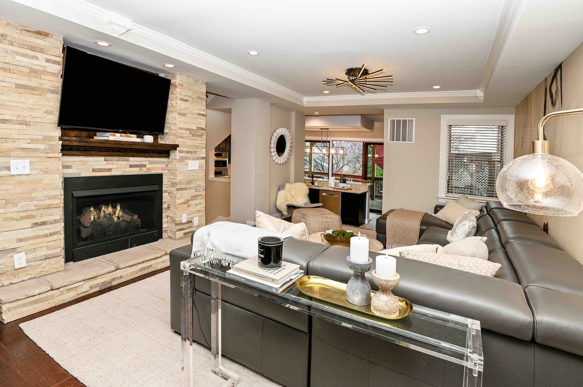 A modern living room features a large sectional sofa arranged around a stone fireplace. A flat-screen TV is mounted above the mantle, while natural light enters through nearby windows. Coffee tables with decorative items sit on a light-colored rug, enhancing the inviting layout.