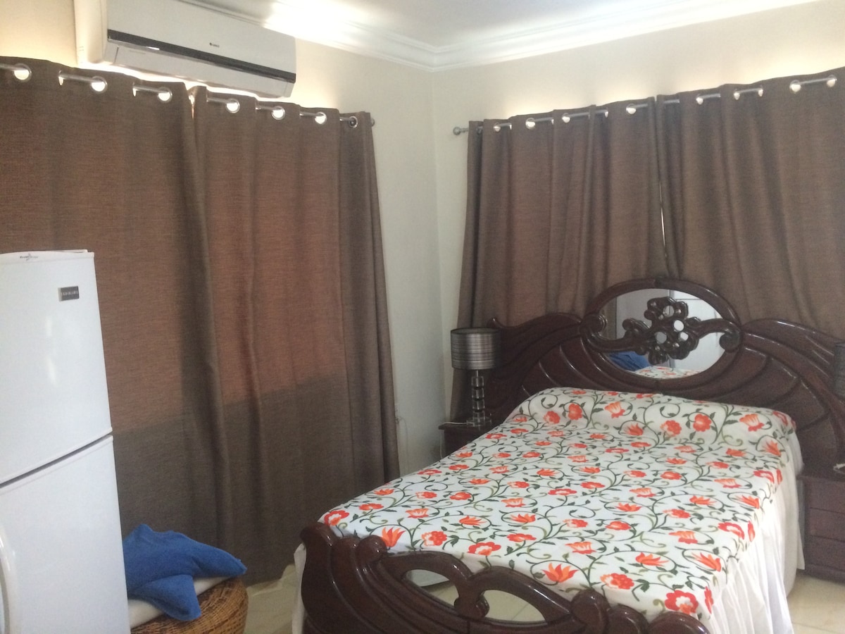 A bedroom features a wooden bed with a floral-patterned bedspread. Brown curtains drape over the windows, allowing soft light to filter in. A small bedside lamp and a refrigerator are visible, adding functionality to the space.