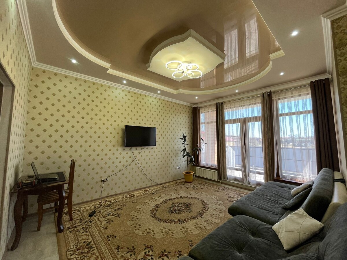 A spacious living area is highlighted by a large, elegant rug on the floor, with a comfortable sectional sofa positioned nearby. Natural light filters through large windows covered with sheer curtains, enhancing the modern ceiling design and wall decor.