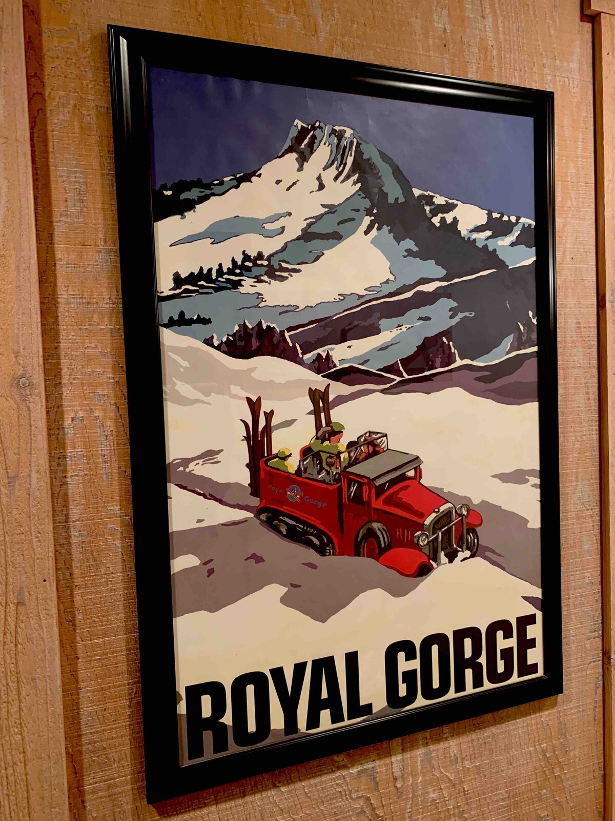 A colorful vintage-style poster depicts a red vehicle equipped for winter, navigating a snowy landscape. Majestic mountains rise in the background, and the title 'ROYAL GORGE' is prominently displayed at the bottom. The artwork is framed and mounted on a wooden wall.