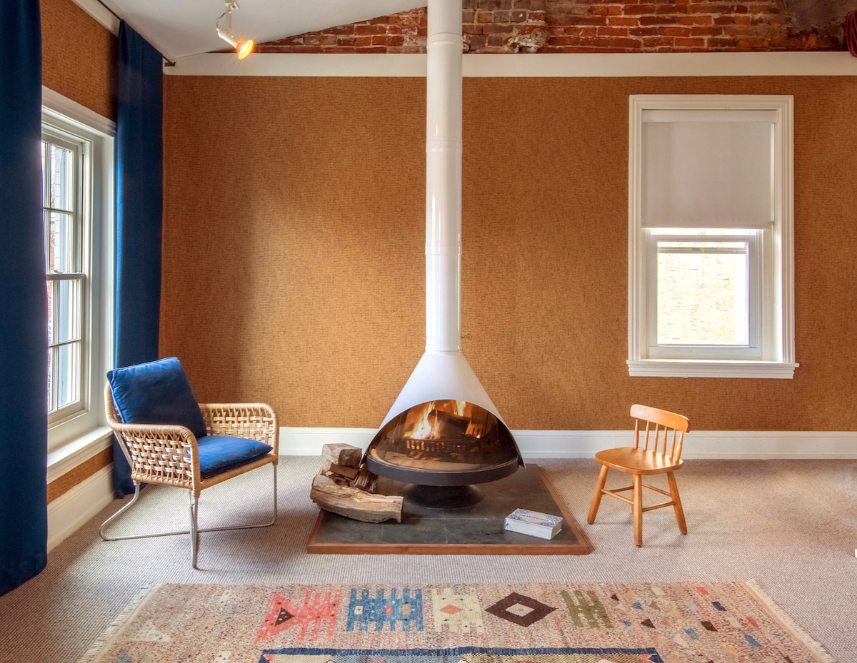 A feature of the room is a modern white fireplace, serving as a focal point on the simple stone hearth. Nearby, a woven chair and a small wooden child's chair are positioned on a colorful area rug, enhancing the cozy ambiance.