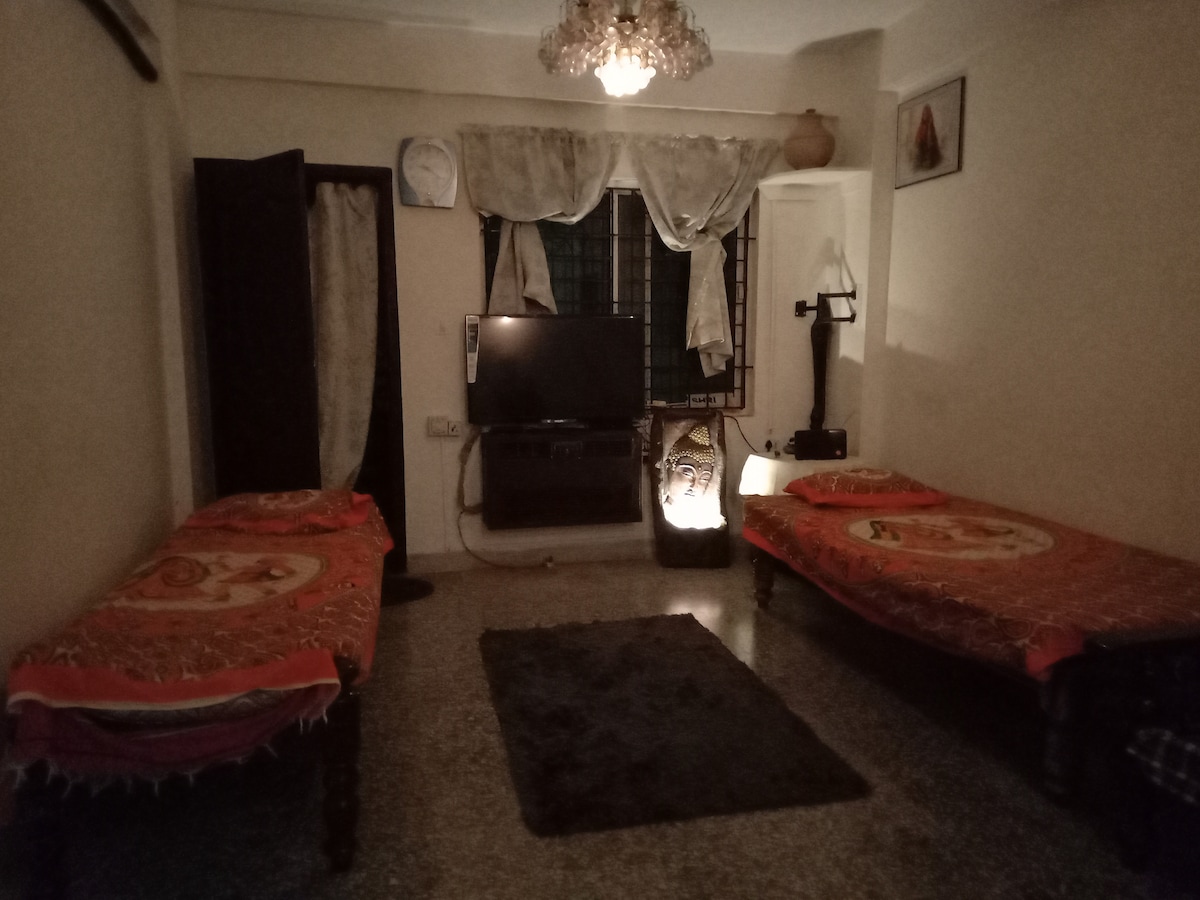 A neatly arranged room contains two beds with colorful covers, positioned opposite each other. A large television is mounted on the wall, facing a soft area rug. Natural light filters through the window, partially covered by sheer curtains.