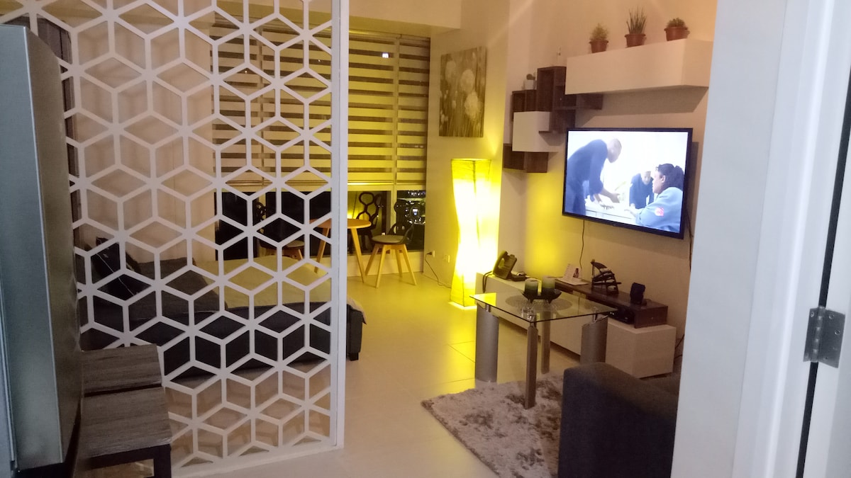 A modern living area features a geometric partition that defines the space. A comfortable sofa is positioned in front of a wall-mounted television. Soft lighting creates a relaxing ambiance, while decorative plants and a stylish side table enhance the minimalist aesthetic.