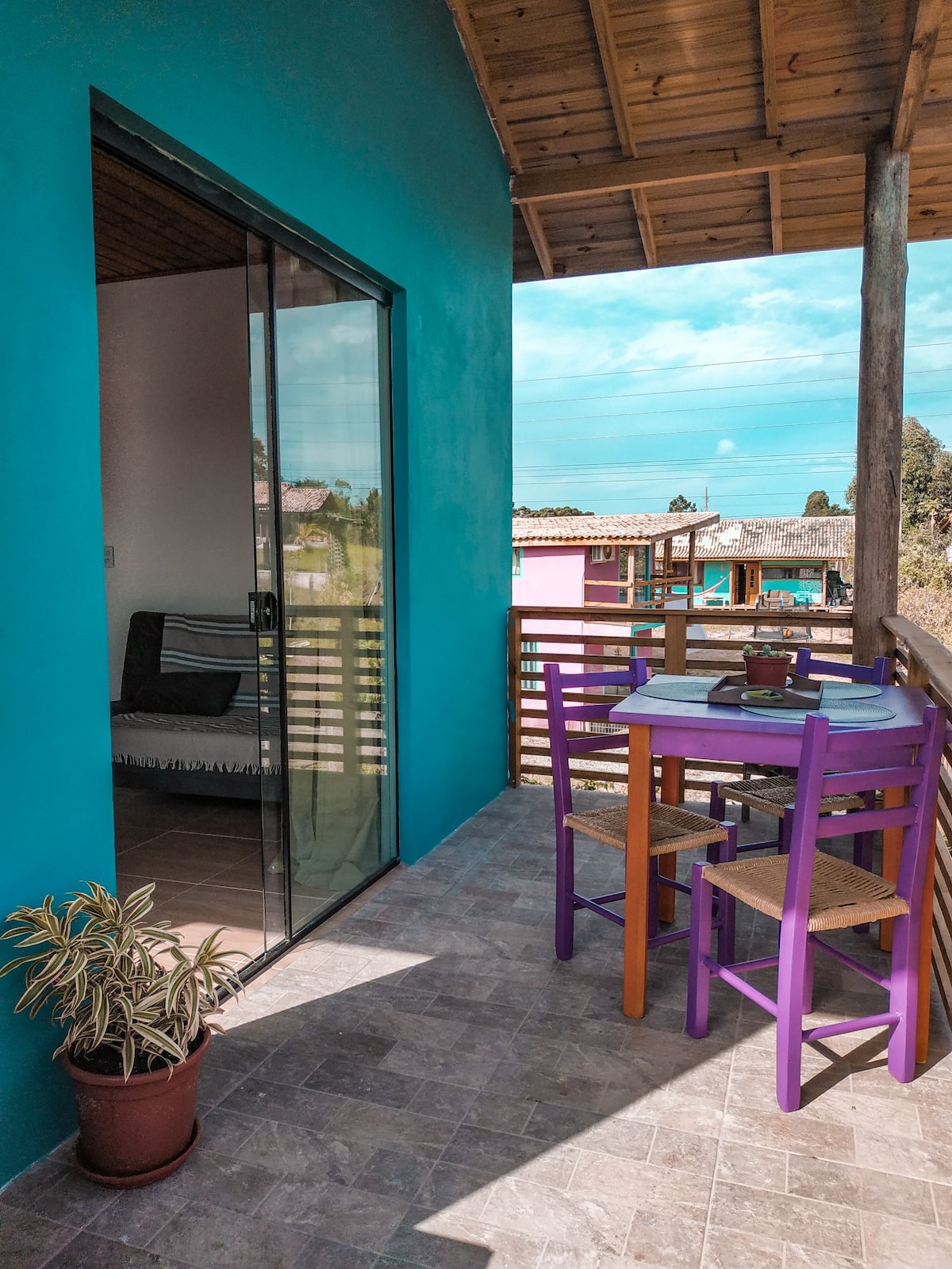 A private balcony features a small dining table set for four, surrounded by colorful chairs. A potted plant is positioned on the side, and sliding glass doors lead into the apartment interior, providing an indoor-outdoor flow and natural light.