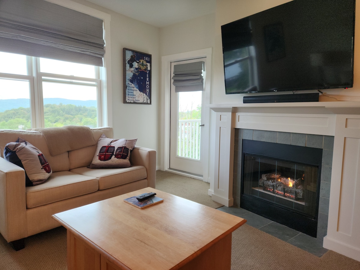 The living area features a comfortable couch and a wooden coffee table. A large flat-screen television is mounted above a stone fireplace, which is lit and creating a soft glow. Natural light enters through the framed window, revealing scenic views of the surrounding landscape.