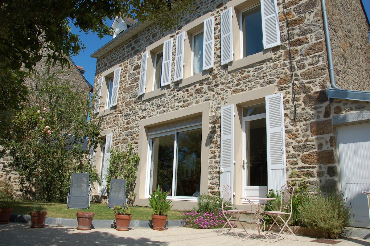 A charming stone house with light-colored shutters stands under a blue sky. Two lounge chairs are positioned on the gravel area in front, surrounded by potted plants. A large glass sliding door allows natural light into the interior.