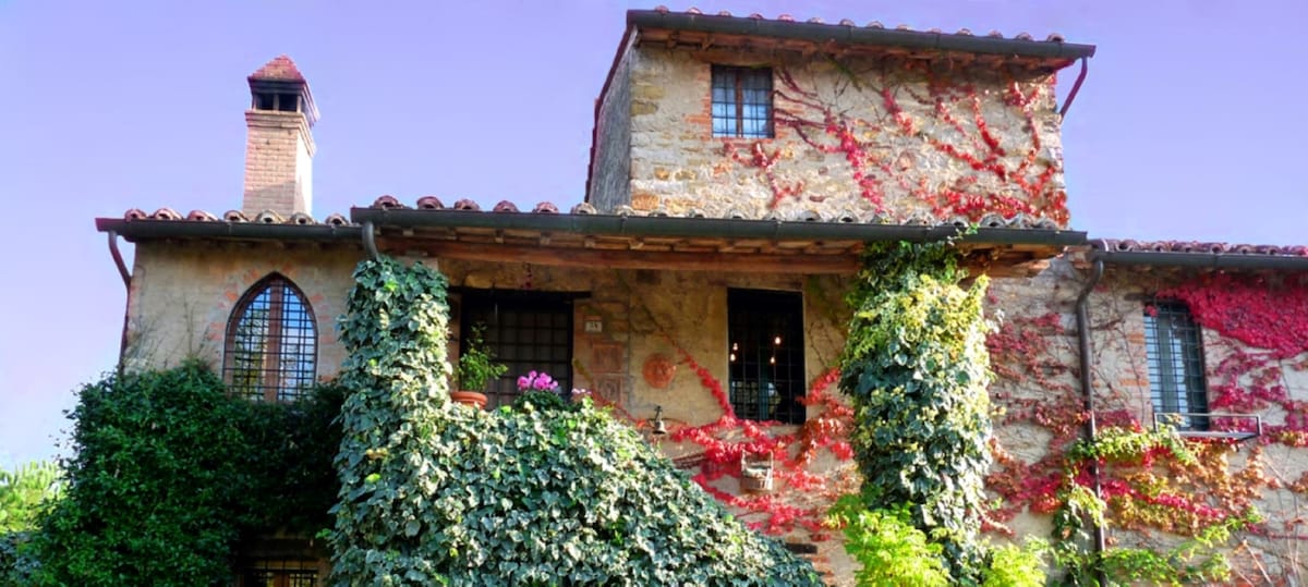 A charming villa exterior is showcased, featuring natural stone walls adorned with vibrant greenery and colorful flowers. The traditional tiled roof and window grilles contribute to its historic character, while bursts of red vines add visual interest against the warm stone.