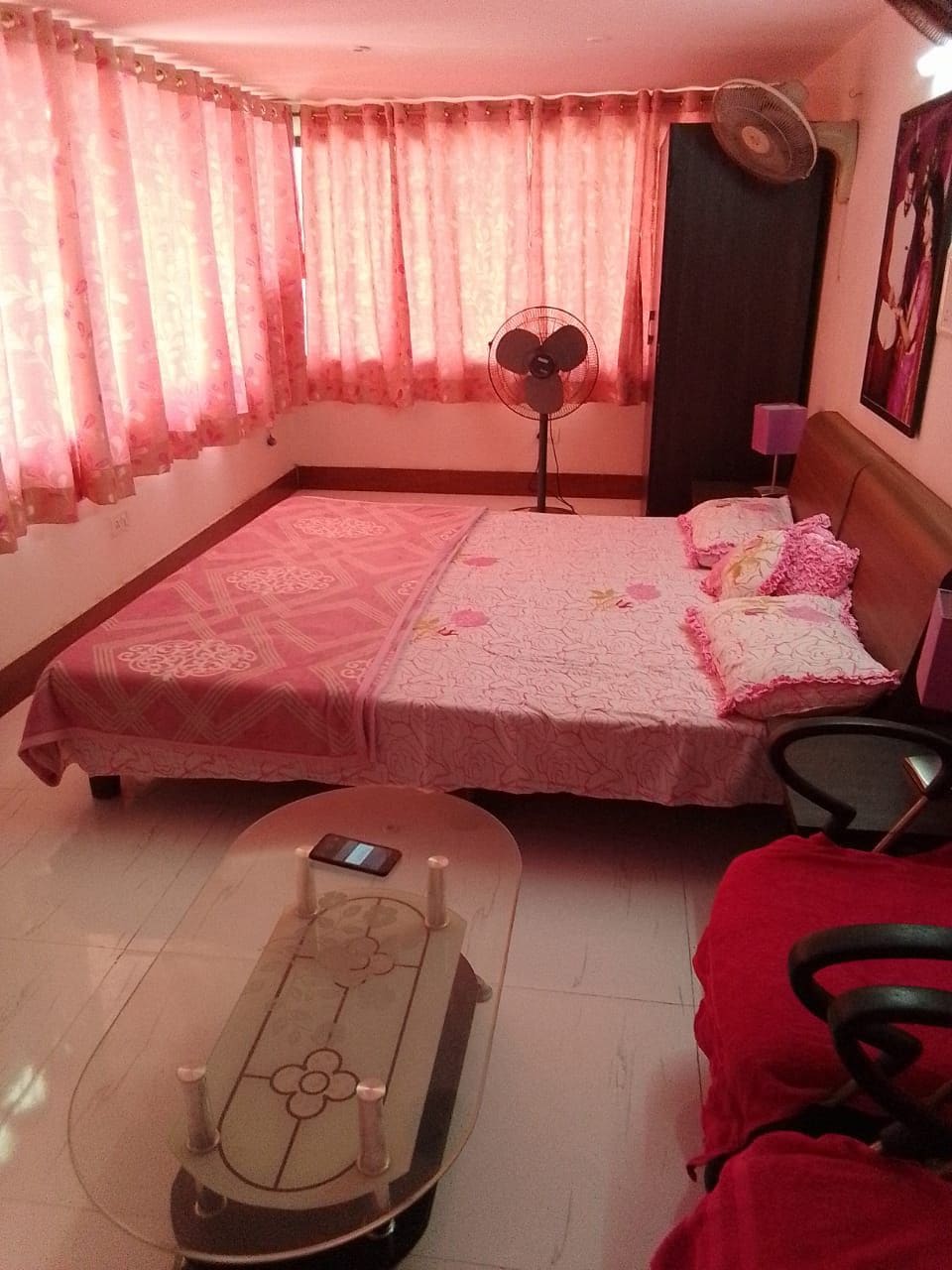 A bright sleeping area features a spacious bed adorned with floral linens. Two large windows, covered with sheer pink curtains, allow natural light to fill the room. A glass coffee table stands in the center, accompanied by a comfortable chair and a standing fan.