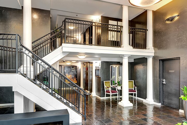 The entrance area features a spacious lobby with polished stone flooring, complemented by decorative columns and a wrought iron staircase. Two chairs and a small table are arranged near the entrance, with potted plants adding a touch of greenery.