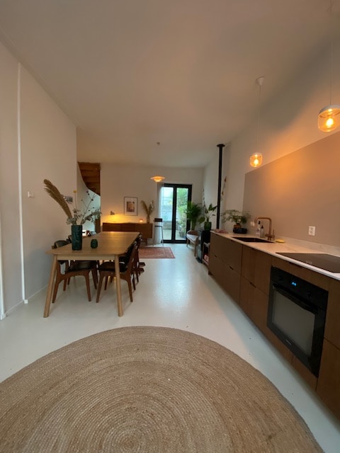 A spacious dining area is visible, featuring a wooden table surrounded by several chairs. Soft lighting is provided by hanging pendant lights. A natural fiber rug is placed on the floor, while a modern kitchen with a white countertop and dark cabinetry is situated in the background.