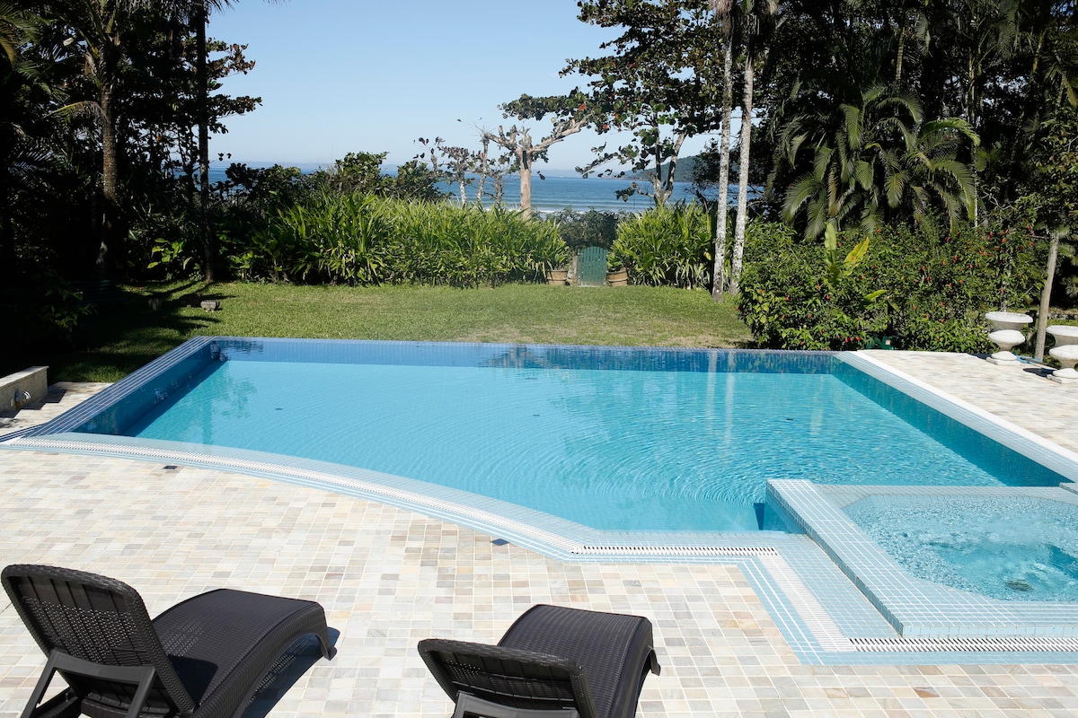 A sparkling pool is positioned beside a relaxing jacuzzi, surrounded by lush greenery and a well-maintained garden. The tranquil ocean can be glimpsed in the background, creating a serene outdoor space ideal for leisure.