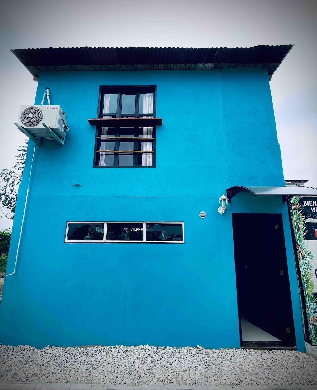 The vibrant blue exterior of the two-story building is complemented by a dark roof and a black entrance door. An air conditioning unit is mounted on the side, while large windows provide natural light. Pebbled ground surrounds the structure, enhancing its inviting appearance.