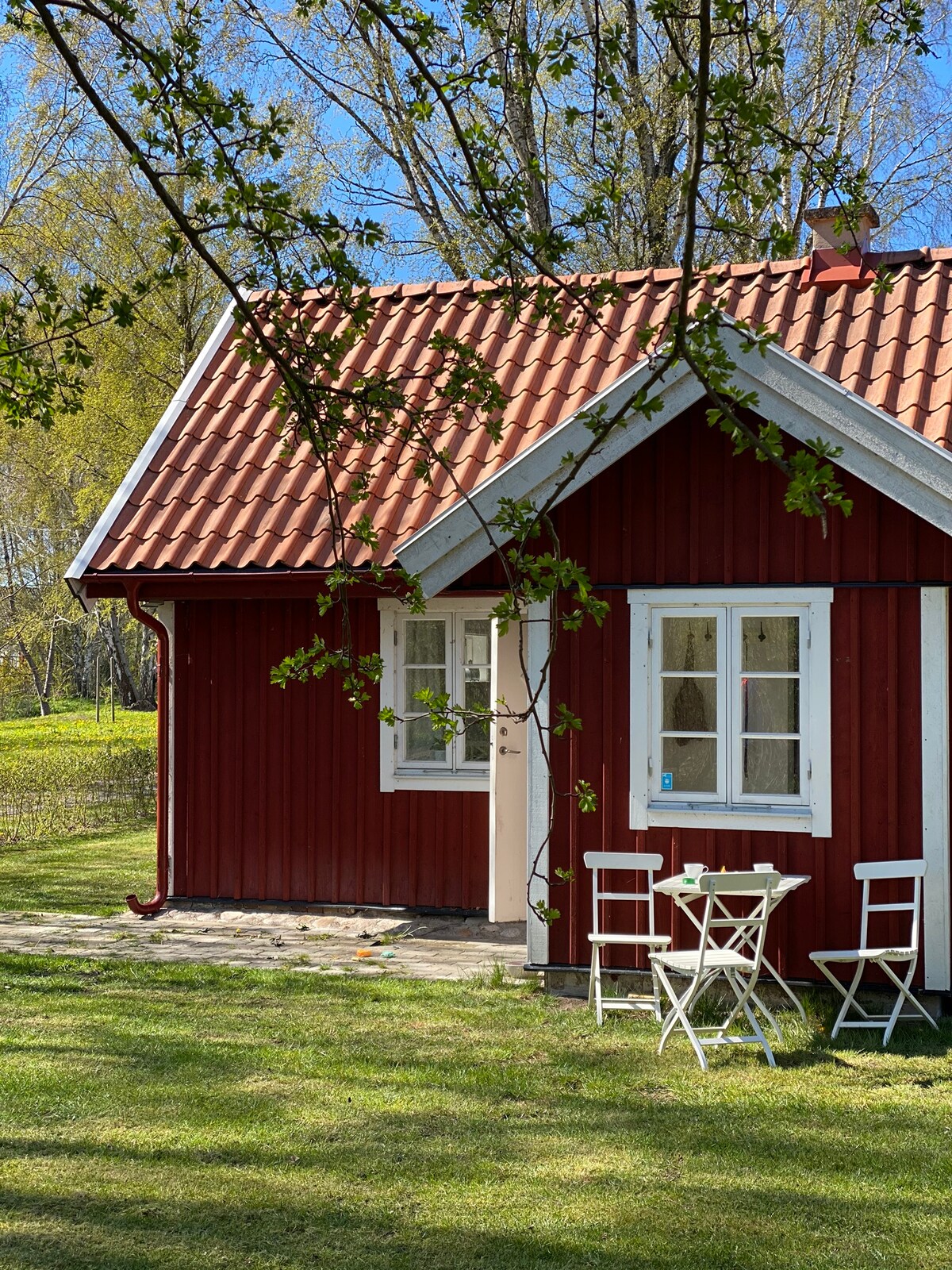 A charming red cottage is depicted, featuring a distinctive red facade and a classic tiled roof. A small outdoor seating area with white chairs is visible, surrounded by trees and lush green grass, creating a peaceful outdoor space.