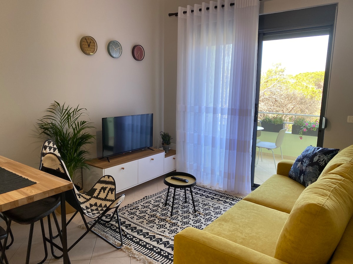 A living area features a yellow sofa complemented by a patterned area rug. A television is positioned on a wooden console beneath decorative wall clocks. Light filters through sheer curtains, revealing an outdoor balcony with a table and chairs, while a potted plant adds greenery.