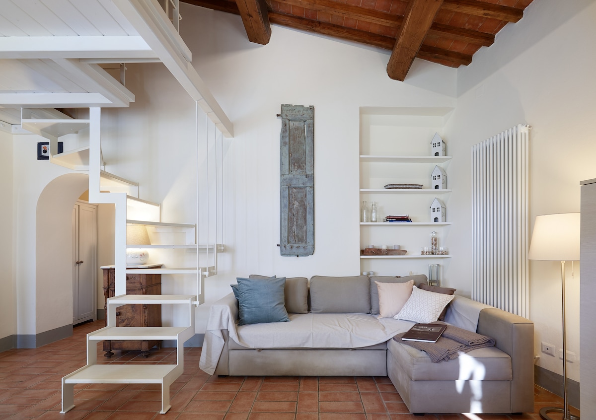A welcoming living area is featured, showcasing a light-toned sofa with neutral cushions. A minimalist staircase leads to the upper level, while shelves display decorative items. Warm light emanates from a nearby lamp, highlighting the natural wooden ceiling beams and terracotta floor tiles.
