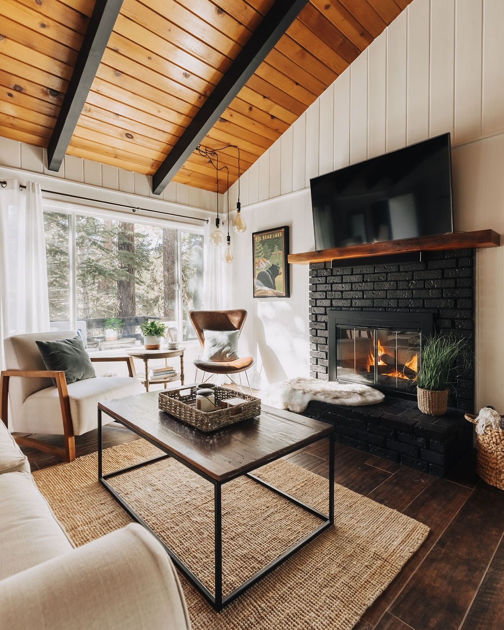 The living area is characterized by wood vaulted ceilings and large windows that allow natural light to fill the space. A gas fireplace is nestled into a black brick surround, while comfortable seating options include a light-colored couch and a stylish armchair. A coffee table sits atop a woven rug.
