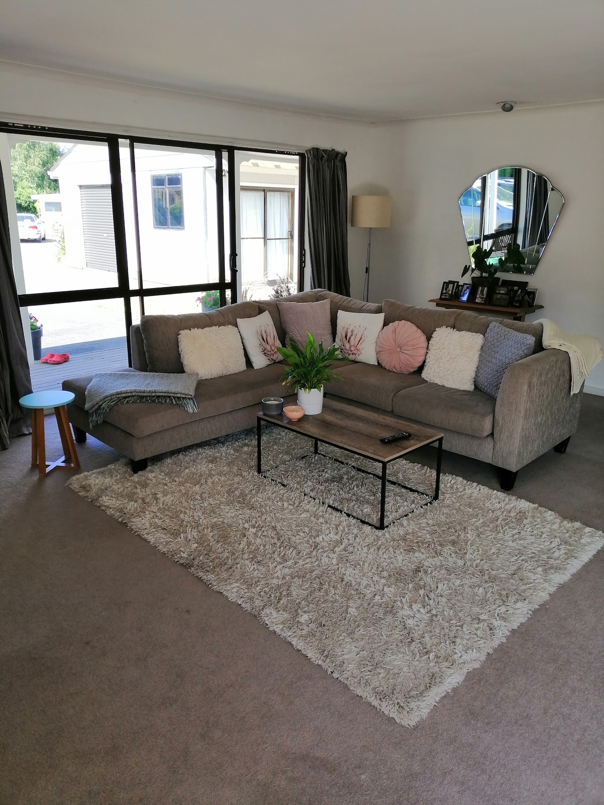 A comfortable living area features a spacious, L-shaped sofa adorned with various cushions. A small side table is positioned beside the sofa, and a coffee table holds decorative items. Light enters through large windows, illuminating the plush carpet and surrounding decor.