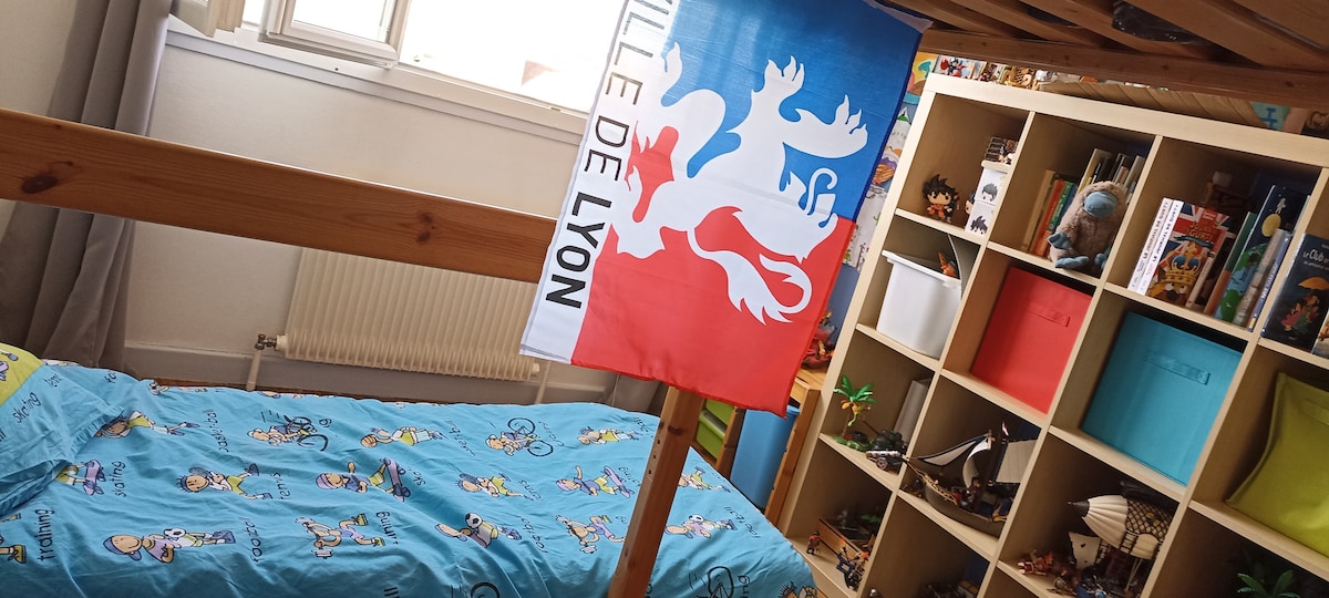 A vibrant children's bedroom features a wooden loft bed adorned with the flag of Lyon. The bedding is decorated with playful patterns. A shelving unit is visible in the background, filled with various toys and colorful storage bins. Natural light streams in from the window.