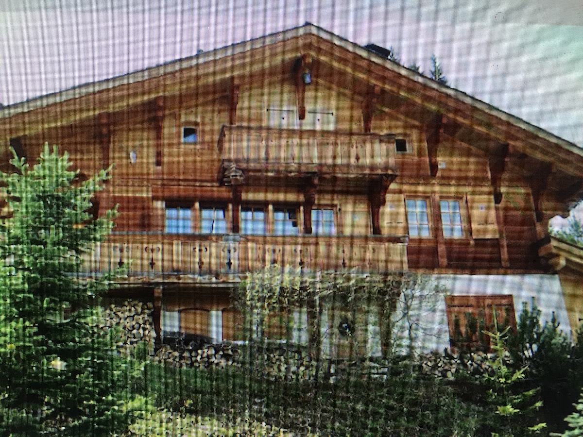 A charming wooden chalet is depicted with a steeply pitched roof and multiple balconies. Natural wood accents are visible along the exterior. Below, stacks of firewood are neatly arranged, and surrounding greenery adds to the serene setting.