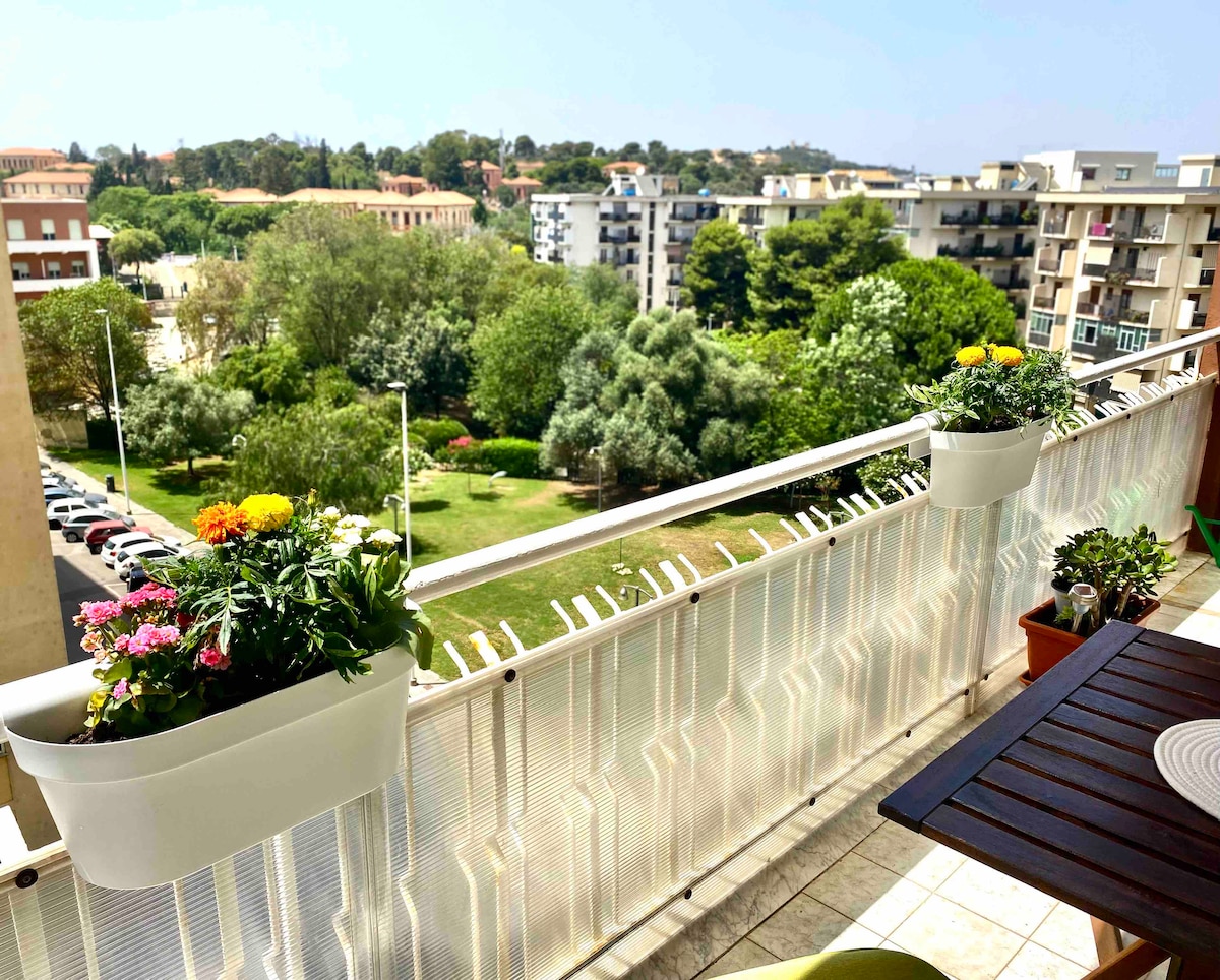 A private balcony displays vibrant flower pots, offering a view of a green park with trees and nearby buildings. The decor includes a wooden table and chairs, inviting relaxation while enjoying the surroundings.