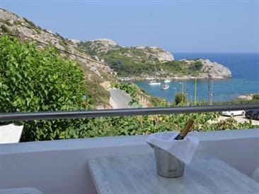 A view of a coastal landscape features rolling hills covered in greenery, leading down to a serene bay with boats anchored in the distance. A small table with a bucket of chilled drinks sits in the foreground, inviting relaxation amidst the picturesque scenery.