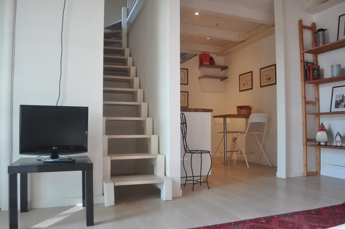 An open living area is shown with a compact television on a small table. A stylish wire chair is positioned nearby. A staircase leads to an upper level, while a kitchen area with bar stools is visible in the background. Natural light illuminates the space.