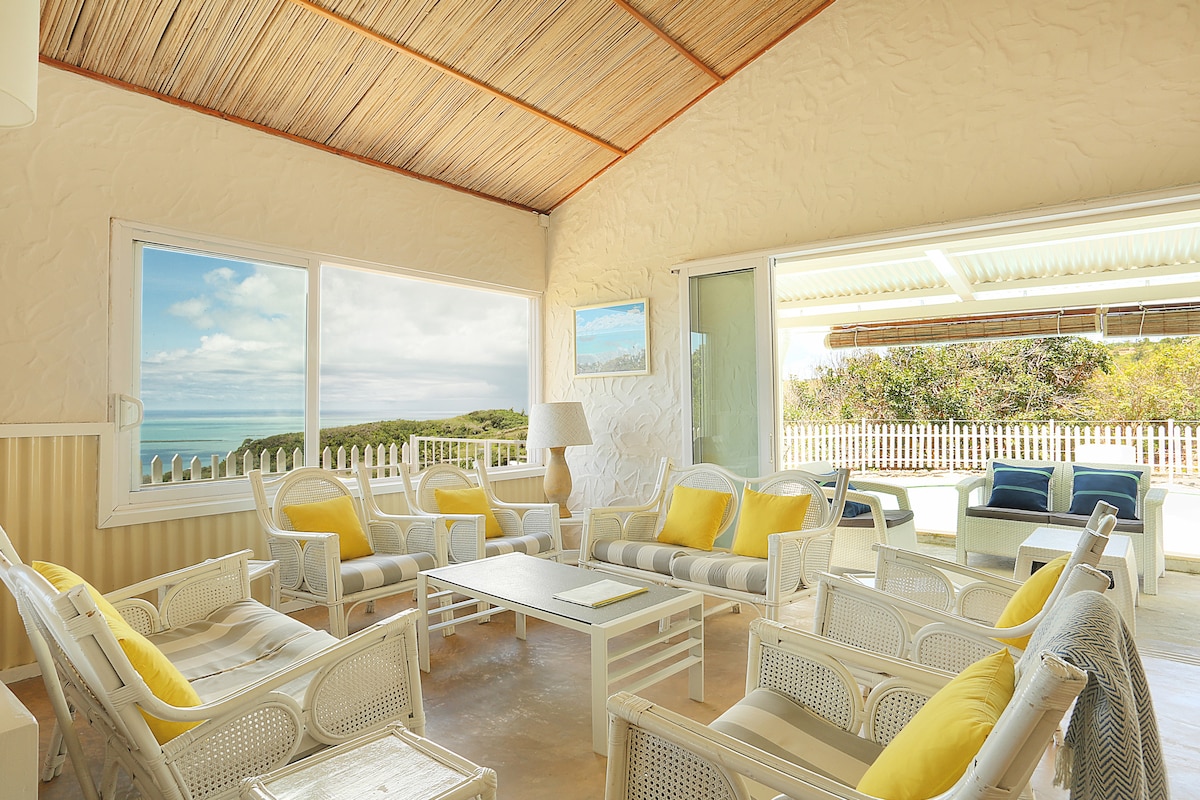 A light-filled living area is showcased with comfortable seating arranged around a central coffee table. Large windows allow for expansive views of the surrounding landscape, highlighting the serene colors of the lagoon. Bright yellow cushions add a refreshing touch to the seating.