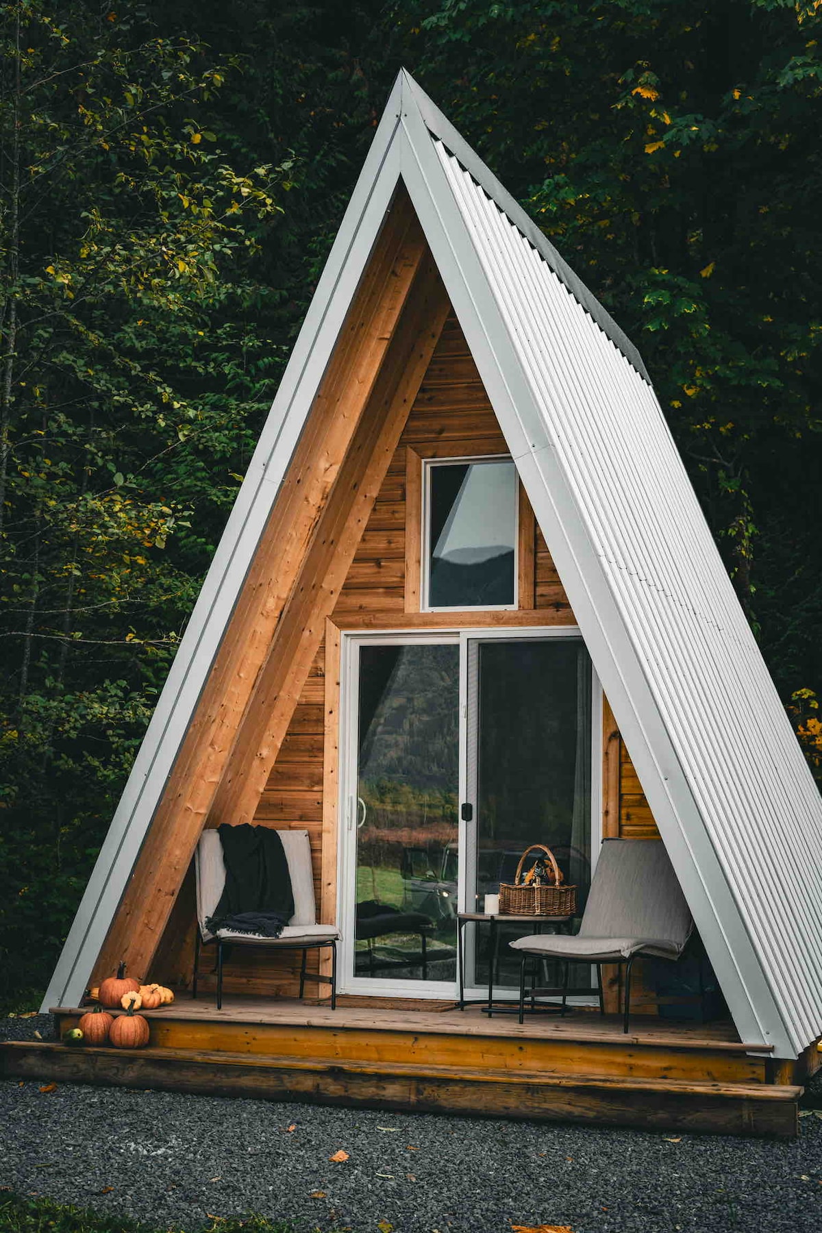 A modern A-frame cabin features a striking triangular structure with wood paneling and a metal roof. A deck is visible with two outdoor chairs and a small table, complemented by decorative pumpkins. Large glass doors provide an inviting entrance, surrounded by lush greenery.