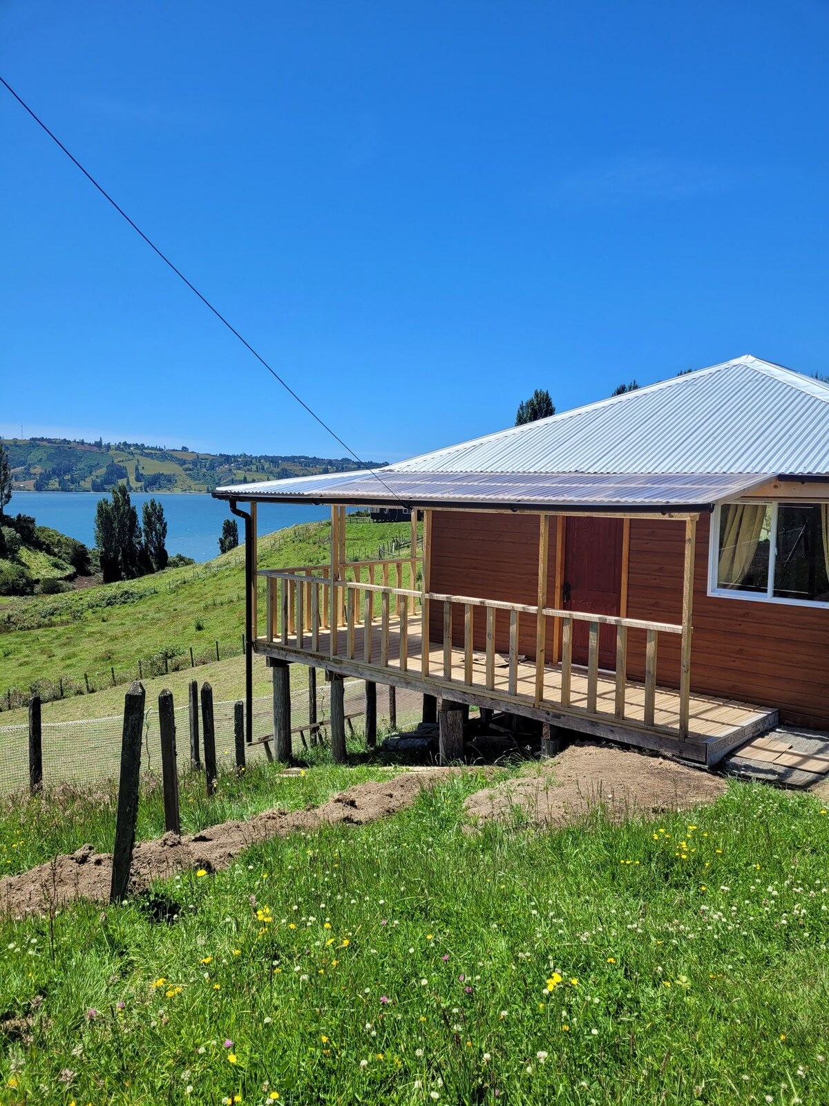 A wooden house is positioned on a hillside, featuring a covered deck supported by wooden railings. The serene landscape includes lush green grass and a clear view of the water in the background, with trees dotting the scenery under a bright blue sky.